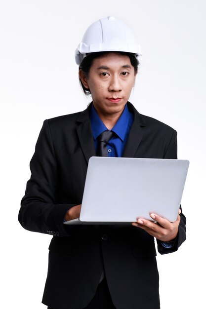 Isolated studio shot of Asian professional successful male foreman industrial engineer in black formal suit and safety helmet standing typing report on laptop notebook computer on white background.