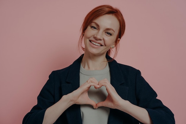 Isolated studio shot of young kind smiling redhead female making heart shape with both hands