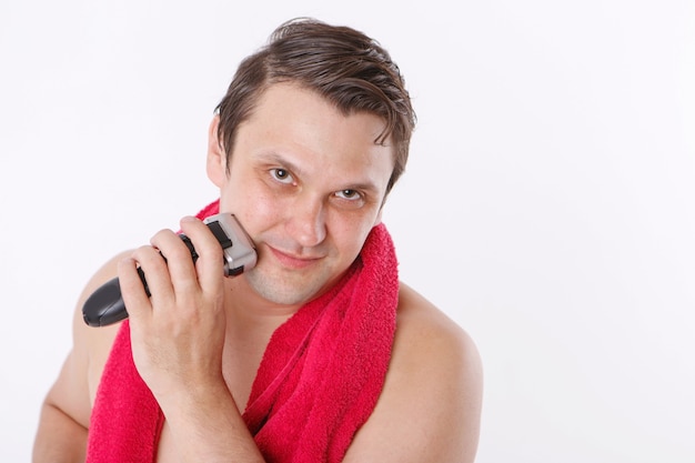 Isolated on a white background: a man shaves his stubble. the guy cleans his beard with an electric razor. morning treatments in the bathroom. red towel around her neck. copy space