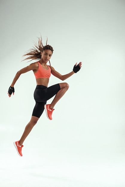 Do it! Full length of young african woman with perfect body in sports clothing jumping in studio against grey background and looking at camera. Sport concept. Motivation. Healthy lifestyle