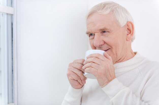 家はとても暖かいです！一杯のコーヒーを持って、窓の近くに立っている間目をそらしている陽気な年配の男性