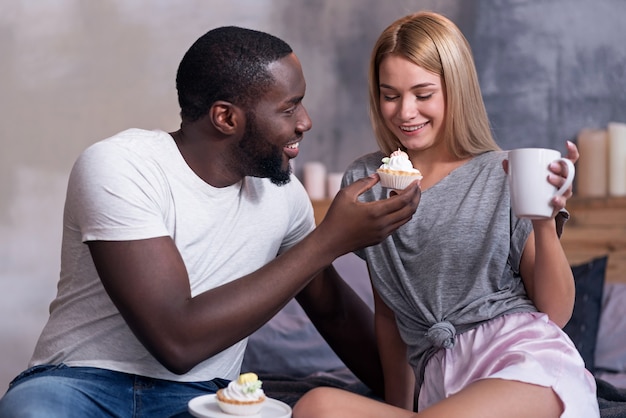 美味しいです。ベッドに座って朝を楽しんでいる間、カップケーキで彼のガールフレンドを養うアフリカ系アメリカ人の若い思慮深い男。