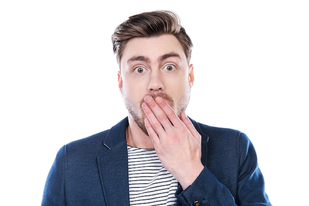 It is unbelievable! Portrait of surprised young man looking at camera and covering mouth with hand while standing against white background