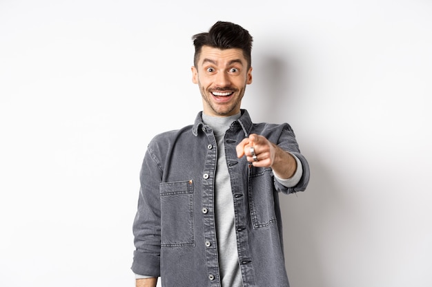 It is you. Excited smiling man pointing fingers at camera with cheerful face, recognize person, standing in denim jacket on white background.