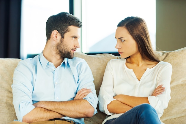 It is your fault! Angry young couple looking at each other and keeping arms crossed while sitting close to each other on the couch