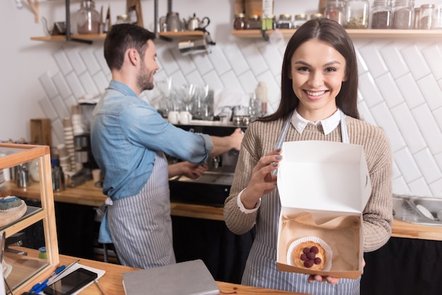 それはあなたを幸せにします。彼女のパートナーがコーヒーを準備している間、笑顔でケーキと箱を保持している美しい幸せな女性。