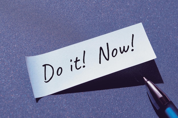 Photo do it now motivator message written on post it note sticky paper with pen single simple minimal motivation note stiker on blue paper background