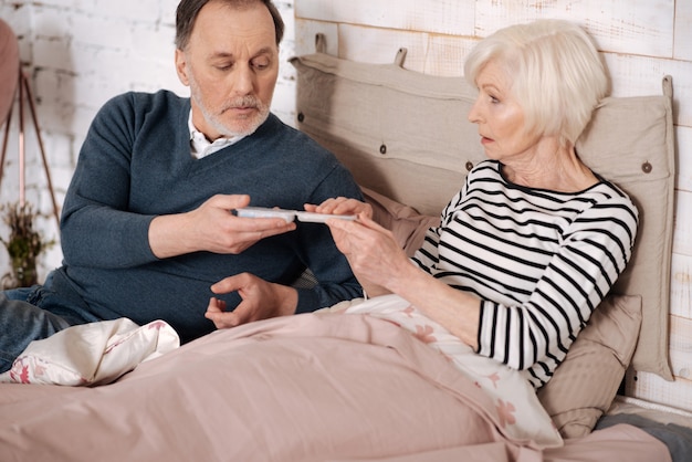 It will help. Senior handsome man is giving pills case to his elderly sick wife lying on bed covered with warm blanket.