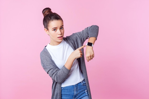 It39s late Portrait of worried brunette teenage girl with bun hairstyle in casual clothes pointing at wristwatches checking time looking displeased impatient studio shot isolated on pink background