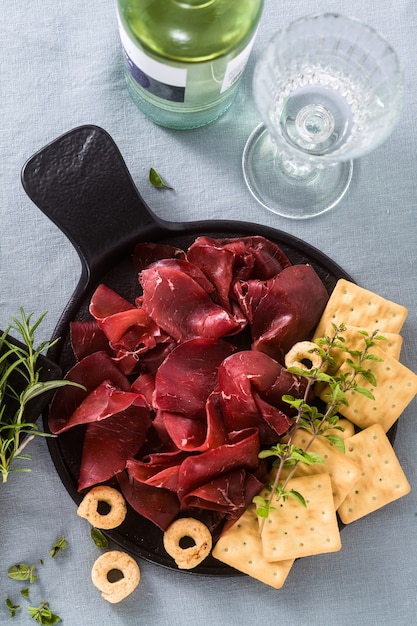 Italiaanse bresaola geserveerd gesneden op een dienblad op een tafel met witte wijn, crackers, grissini en taralli met aromatische kruiden op een blauw linnen feestelijk tafelkleed.