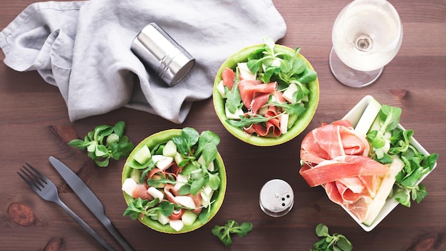 Italiaanse of spaanse salade verse meloen met prosciutto of jamon mozzarella kaas en groene kruiden in meloen plaat witte wijn glas op donkere houten tafelblad weergave mediterrane keuken schotel