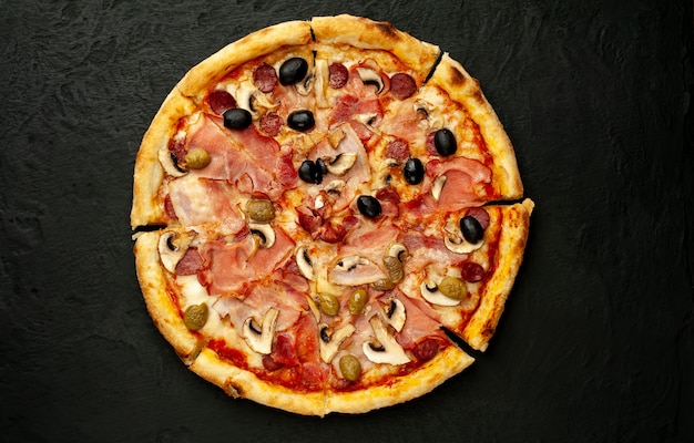 Italiaanse pizza met spek, champignons, olijven, tomaten op een zwarte betonnen achtergrond