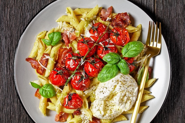 Italian caprese pasta bake of pasta penne, bacon, cherry tomatoes, and mozzarella cheese, topped with shredded parmesan, fresh basil served on a plate on a dark wooden table, top view, close-up