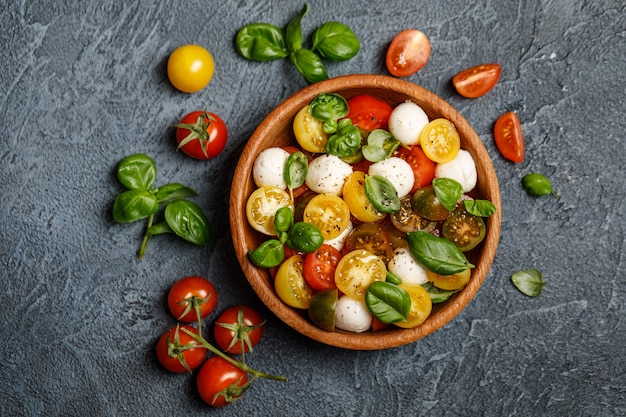 Italian caprese salad with sliced tomatoes, mozzarella cheese, basil, olive oil in wooden bowl.