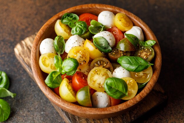 Italian caprese salad with sliced tomatoes, mozzarella cheese, basil, olive oil in wooden bowl.