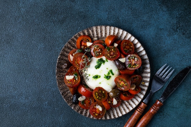 Italian caprese salad with sliced tomatoes, mozzarella cheese, olive oil on dark background. Top view. Black tomato salad with burrata and garlic. Comfort food. Sustainable consumption.