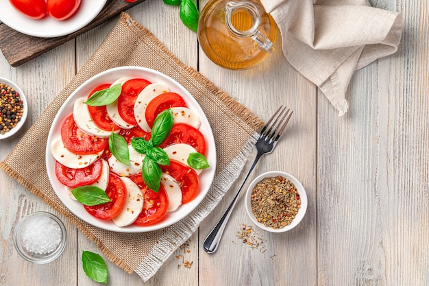 Итальянский салат капрезе с помидорами, сыром моцарелла и базиликом в тарелке на бежевом деревянном фоне. Здоровое, диетическое питание.