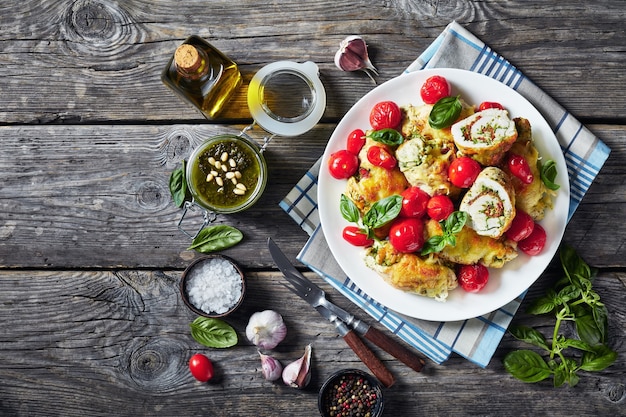 Фото Итальянские куриные рулетики с соусом песто, плавленым сыром и помидорами на белой тарелке с ингредиентами на деревянном столе, горизонтальный вид сверху, плоская планировка, копировальное пространство