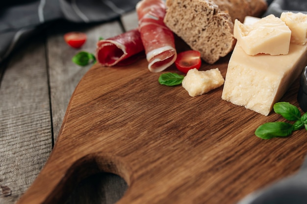 Italian cuisine concept. Cutting board, Parmesan Cheese chunks, thinly sliced ham, cuts of fresh cherry tomatos, basil leaves branches, crusty bread toast, kitchen towel on wooden table