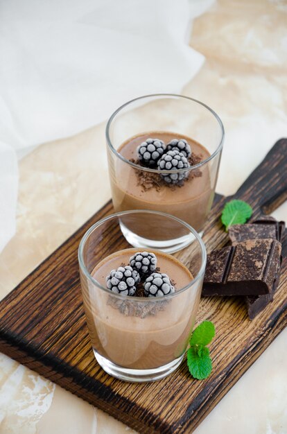 Italian dessert - chocolate panna cotta, mousse, cream or pudding with blackberry in a glass on a board on a light concrete background. Vertical orientation. Copy space.