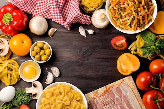 Italian food ingredients with pasta, vegetables, mushrooms, olives and bacon. Flat lay of mediterranean cuisine ingredients on dark wooden background, copy space