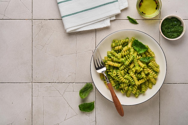 イタリアのフジッリパスタとペストフジッリパスタとバジルペストとハーブパルメザンチーズバジルとニンニクの白いプレートに古いひびの入ったタイルテーブルコンクリートの背景上面図モックアップ