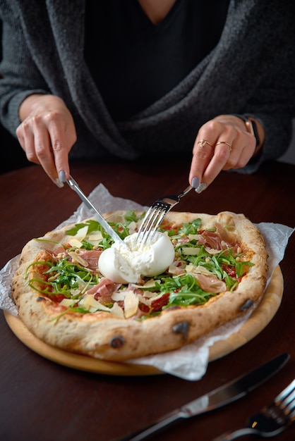 Italian pizza restaurant menu - pizza met burrata, prosciutto en rucola, met vrouwelijke handen in het frame.