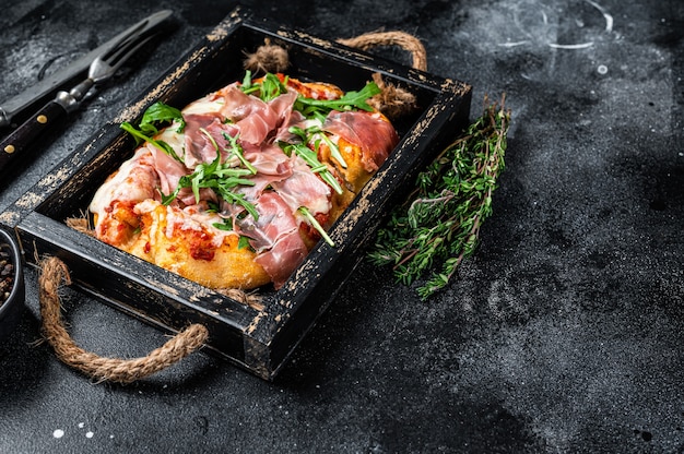 Italian Pizza with prosciutto parma ham, arugula salad and cheese in a rustic wooden tray. Black background. Top view. Copy space.