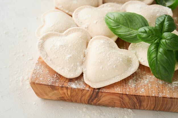 Italian ravioli pasta in heart shape Tasty raw ravioli with flour and basil on white background Food cooking ingredients background Valentines or Mothers Day lunch ideas Top view with copy space