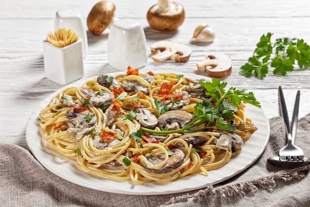 Italian spaghetti with cremini mushroom sauce of heavy cream and parmesan cheese with crispy bacon and parsley served on a white plate and cutlery on a white wooden background, top view, close-up