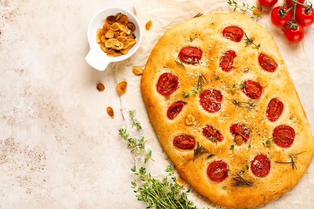 Italian traditional focaccia bread baking with with cherry tomatoes, parmesan and rosemary on light brown background. Top view
