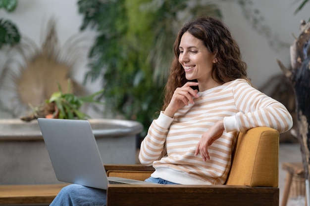 Italian woman waving hello having video call on laptop sitting in armchair in cozy home garden