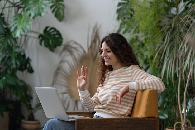 Italian woman waving hello having video call on laptop sitting in armchair in cozy home garden