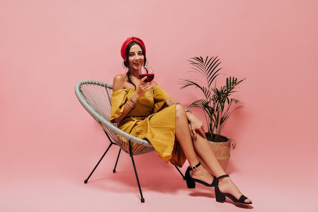 Italian woman with curly hair in round earrings red headband yellow dress and stylish shoes looking into camera smiling and holding glass of wine