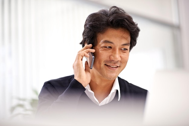 Это один из тех напряженных дней в офисе. Снимок красивого азиатского бизнесмена, разговаривающего по телефону во время работы за своим столом.