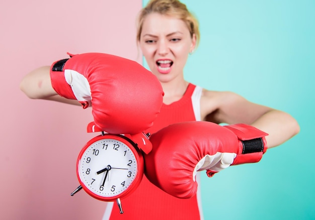 Пришло время для тренировки по боксу Эмоциональная женщина держит часы в боксерских перчатках Милый боксер с сердитым взглядом борется с будильником Спортсменка бьет часы На часах еще есть время