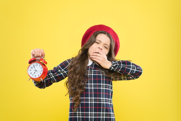 Its time to go to sleep. Sleepy little girl in evening time on yellow background. Yawning small child holding alarm clock. Late time. Break time, refresh yourself.