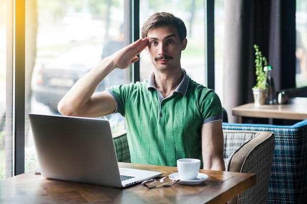 Ja meneer Jonge serieuze zakenman in groene tshirt zitten en kijken naar camera met saluut gebaar en klaar om de missie bedrijfsconcept binnen schot in de buurt van groot raam overdag te voltooien