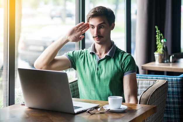 Ja meneer jonge zakenman in groen t-shirt zittend en kijkend naar laptopscherm op videogesprek met saluutgebaar en klaar om de missie te voltooien, binnenbedrijf dat overdag in de buurt van groot raam is geschoten