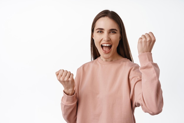 Ja winnaar, laten we het vieren. Glimlachend opgewonden meisje dat handen omhoog steekt, vuist pompt en van tevredenheid schreeuwt, doel bereikt, triomfeert, wint, staande tegen een witte achtergrond.