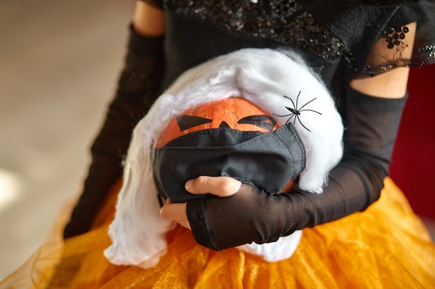 Jack o lantern, halloween-pompoen in medische bescherming zwart masker in meisjeshanden tijdens Halloween-viering thuis covid19 coronavirus pandemie, kopieer ruimte.