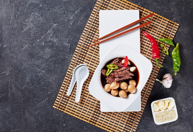 Jangjorim, beef braised in soy sauce served in white bowl with quail eggs, garlic cloves and chili pepper slices, view from above, flatlay, copy space