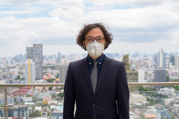 도시 전망에 대한 코로나 바이러스 발발로부터 보호하기 위해 마스크를 쓰고있는 양복에 곱슬 머리를 가진 일본 사업가