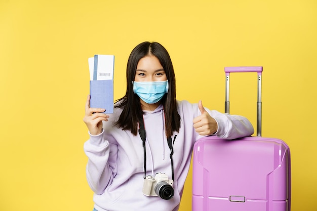 Japanese girl in medical mask, showing her passport and flight travel pass tickets, thumbs up, posing with suitcase, going on vacation, yellow background