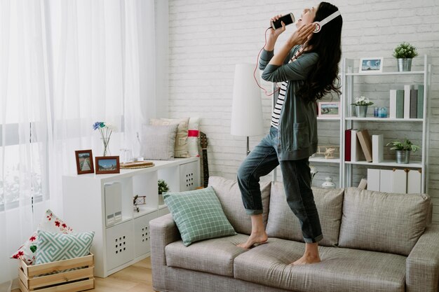 japanese girl standing on sofa wearing earphones is utterly absorbed in singing. relaxing asian young woman is chanting loudly using mobile as microphone.