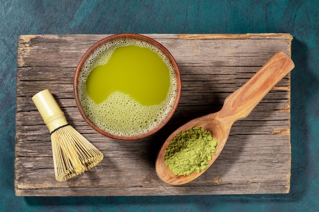 Japanese green matcha tea in wooden cup, matcha powder in wooden spoon and bamboo whisk on old wooden board. Top view.