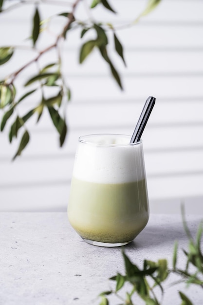 Japanese matcha latte green tea with soy milk and spoon in glass on light background with branches and shadow