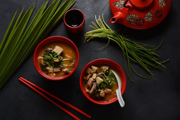 Японский суп мисо в двух красных японских мисках на черном столе. Мисо-суп с тофу, тунцом, зеленью и водорослями с белой японской суповой ложкой и красными палочками для еды.