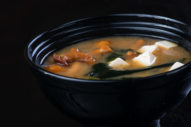 Японский суп с курицей, яйцом, грибами симэдзи и баклажанами на темной стене в черной тарелке.