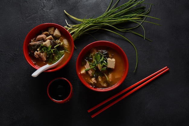 Japanse miso-soep in twee rode Japanse kommen op de zwarte tafel. Misosoep met tofu, tonijn, kruiden en zeewier met witte Japanse soeplepel en rode eetstokjes.